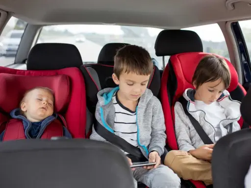 Dziecko śpi w foteliku, a dwaj starsi bracia grają na telefonach. Od kiedy można zapinać dziecko pasami samochodowymi? Od urodzenia, w odpowiednim foteliku.