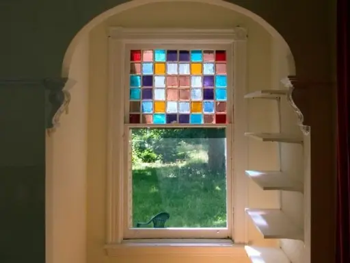 Un vitrail design aux carrés colorés illumine une fenêtre, projetant des reflets joyeux sur l'herbe verdoyante à l'extérieur.