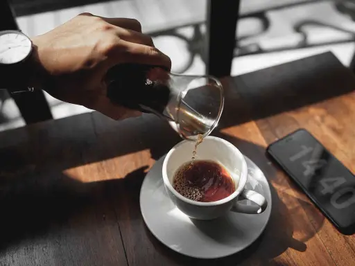 Hand gießt Kaffee in eine Tasse, daneben ein Smartphone mit Uhrzeit.