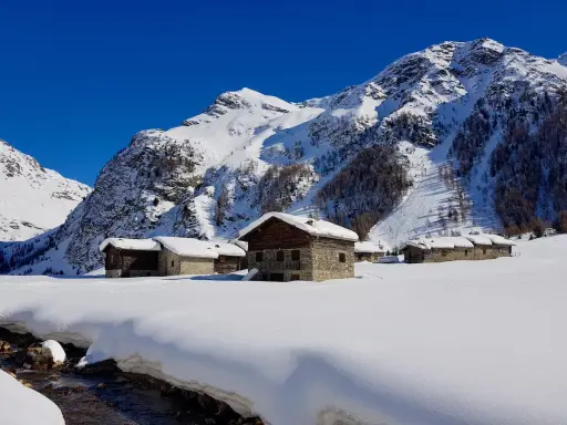 Case innevate nel Val di Rezzalo, con baite in pietra e legno. Per sapere val di rezzalo come arrivare, segui il ruscello che scorre tra la neve.