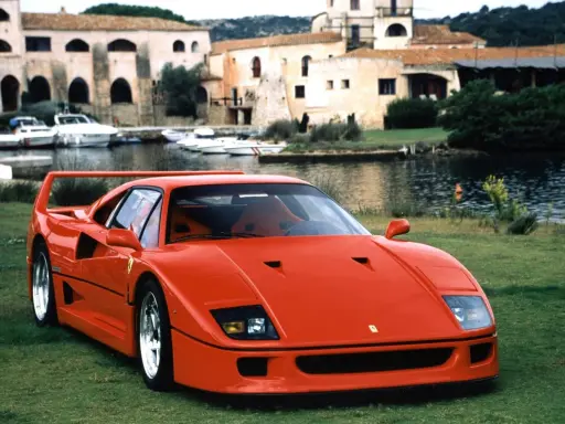 Roter Ferrari F40 auf einer Wiese, im Hintergrund ein Hafen mit Booten und Häusern. Der Ferrari F40 Neupreis war damals ein Traum.
