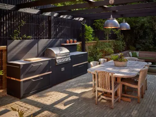 Nowoczesny grill w ogrodzie, z czarną zabudową i drewnianym stołem, zaprasza do wspólnego biesiadowania.