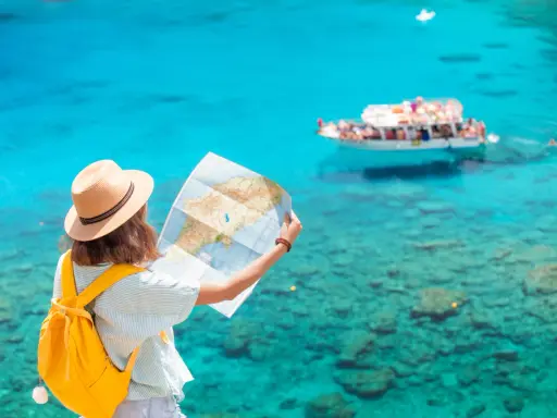 Ragazza con zaino giallo e cappello di paglia studia una mappa, pensando a quanti soldi portare in crociera, con mare turchese e barca sullo sfondo.
