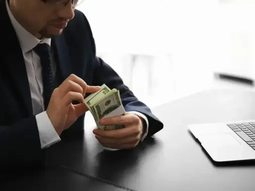 Mężczyzna w garniturze liczy pieniądze, zastanawiając się, ile zarabia osoba pracująca w banku. Obok leży laptop.