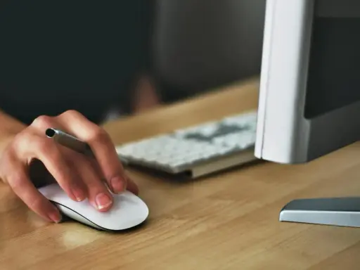 Main souris blanche sur un bureau en bois, avec un clavier et un écran d'ordinateur flous en arrière-plan.