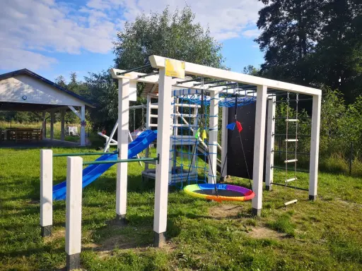Plac zabaw w **dolinie roztoczy**: zjeżdżalnia, drabinki, huśtawka i ścianka wspinaczkowa. W tle altana.
