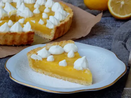 Une part de tarte au citron, garnie de meringue, sur une assiette. La tarte entière est visible en arrière-plan. Une recette de tarte au citron parfaite pour le dessert.