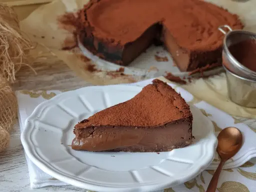 Una porción de tarta de chocolate, cubierta de cacao en polvo, reposa en un plato blanco. El resto de la tarta, también espolvoreada, se ve al fondo.
