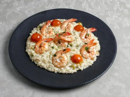 Kremowe risotto z krewetkami, pomidorkami koktajlowymi i posiekaną natką pietruszki na czarnym talerzu.