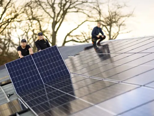 Arbetare installerar solpaneler på ett tak. En av dem, en kvinna, ler glatt medan hon håller i en panel. En annan man hjälper till, och en tredje person sitter på taket. De är en del av teamet för **solverkets alfa**.