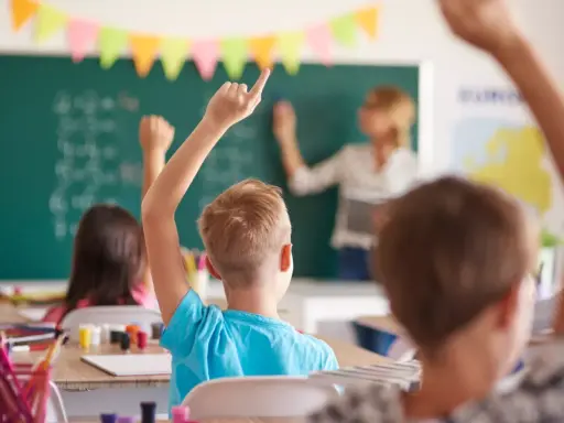 Uczniowie z podniesionymi rękami na lekcji. Nauczycielka pisze na tablicy. To obrazek, który mógłby znaleźć się na karcie nauczyciela.