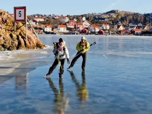 Två personer åker skridskor på en isbelagd sjö, med en pittoresk by i bakgrunden. De använder stavar för att hjälpa sig fram, vilket visar hur åker man skridskor på ett effektivt sätt.