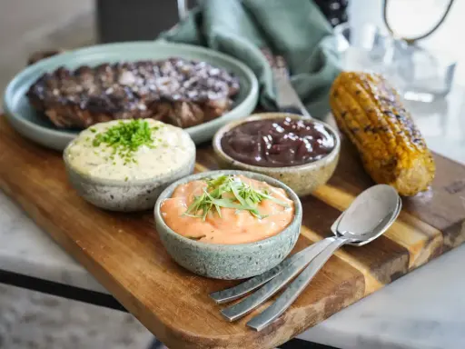 Grillad biff med tre olika såser till grillat: en vit med örter, en mörk och en orange. Majskolv och bestick ligger på en träbricka.