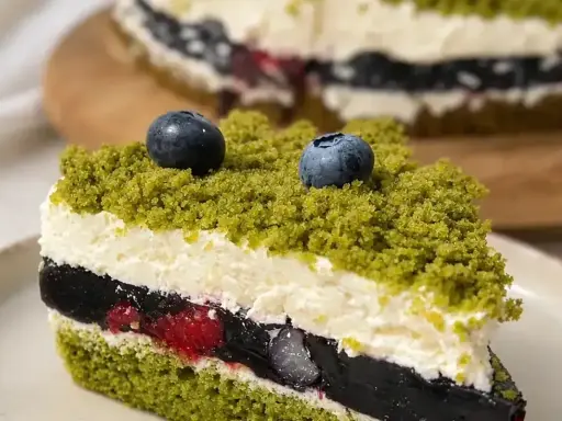 Kawałek tortu z zielonym biszkoptem, kremem, galaretką i owocami, posypany okruszkami przypominającymi leśny mech.
