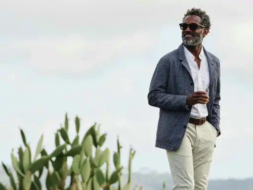 Man in stylish beach wedding attire, wearing a seersucker jacket, white shirt, and light pants, holding a drink.