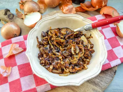 Schüssel mit karamellisierten Zwiebeln, die auf einem karierten Tuch liegen. Ganze und halbierte Zwiebeln sind daneben zu sehen.