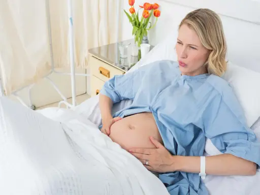 Gravid kvinna i sjukhussäng, med händerna på magen, ser ut att ha värkar. Vattnet har gått och hon väntar på barnet.