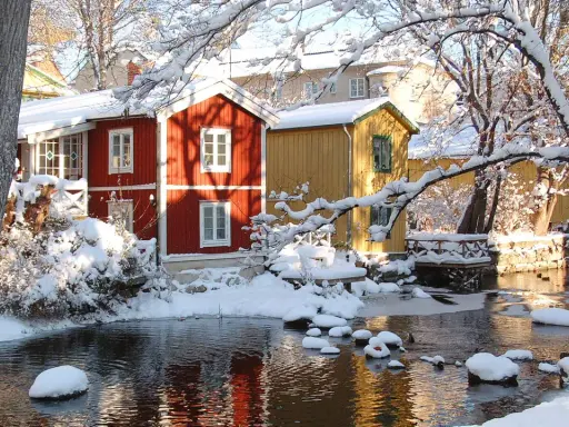 Vinteridyll vid vattnet i destination Gåsvik. Röda och gula hus speglas i det isiga vattnet, omgivna av snötyngda träd.