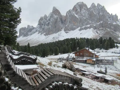Panorami innevati con il rifugio delle Odle in lontananza, panchine in legno e pini.