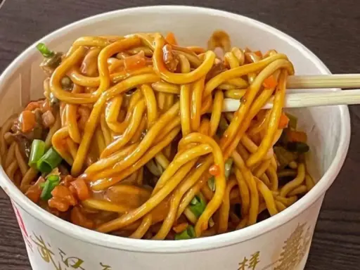 Un tazón lleno de fideos chinos salteados con verduras y salsa. Palillos levantan un bocado.