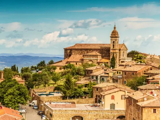 Panoramica del centro storico di Montalcino, con la chiesa e le case in pietra sotto un cielo azzurro con nuvole.