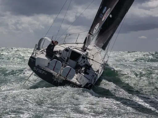 Xavier Macaire navigue sur un voilier dans une mer agitée, le bateau est incliné, les vagues déferlent.