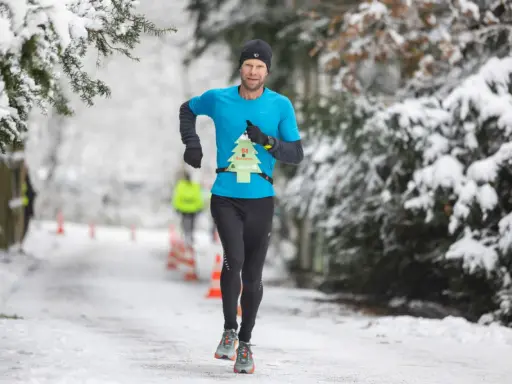 Mann läuft im Winter durch verschneiten Park. Er trägt ein blaues Shirt mit einer Startnummer in Form eines Weihnachtsbaums.