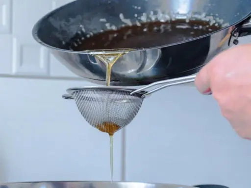 Se cuela aceite rancio de una sartén a través de un colador fino, cayendo en un recipiente.