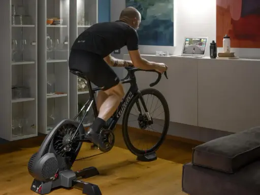 Mężczyzna trenuje na rowerze stacjonarnym z napędem **direct drive**, śledząc wirtualną trasę na laptopie.
