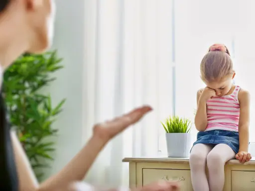 Smutna dziewczynka siedzi przy oknie, a jej mama gestykuluje. Warto nauczyć się, jak przestać krzyczeć na dziecko, by budować lepsze relacje.