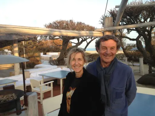 Patrice de Colmont et une femme souriants sur une terrasse de plage, avec des arbres dénudés et des sièges blancs en arrière-plan.