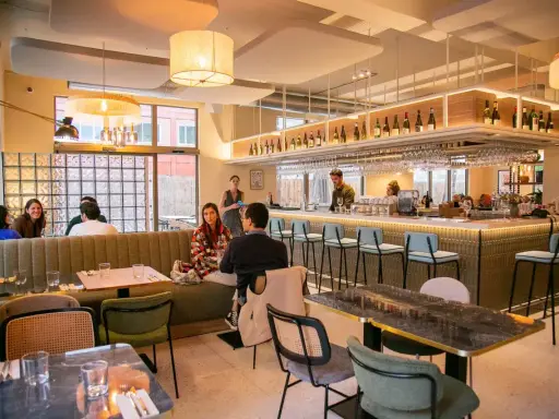 Ambiance chaleureuse d'un restaurant, avec des clients attablés et un bar bien garni. L'esprit du terroir se retrouve dans la sélection des vins et la convivialité.