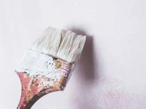 Pincel desgastado con restos de pintura blanca a punto de tocar una pared de cal con textura sutil.
