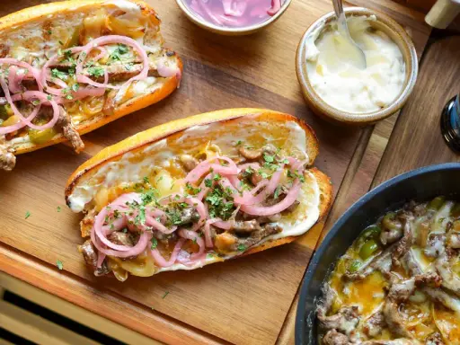 Två philly cheese steak-mackor med möra strimlor av kött, smält ost, lök och paprika, toppade med picklad rödlök och persilja.