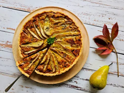Apetyczna tarta z gruszkami, udekorowana natką pietruszki i skórką cytrynową, prezentuje się na drewnianej desce. Obok leży świeża gruszka i jesienne liście.