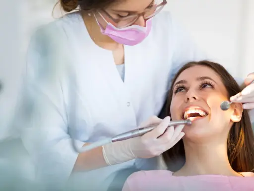 Dentystka w masce i rękawiczkach bada zęby pacjentki za pomocą lusterka i wiertła.