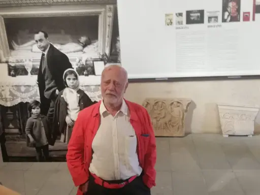 Un uomo anziano con giacca rossa, davanti a una foto di Ferdinando Scianna in Sicilia con bambini.