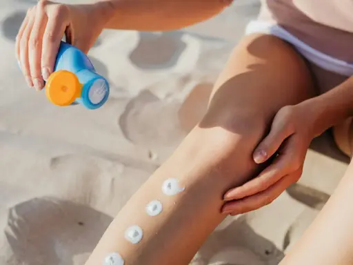 Main sur le sable, une main applique des points de crème solaire sur une jambe. Attention, cette crème solaire perimee pourrait ne plus protéger efficacement.
