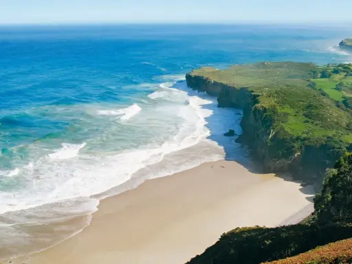 Atemberaubende Küstenlandschaft am Camino del Norte. Grüne Hügel treffen auf das azurblaue Meer mit sanften Wellen, die auf einen einsamen Sandstrand treffen.