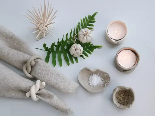 Två ljusstakar av lufttorkande lera med värmeljus, salt- och pepparskålar, servettringar och en dekoration av torkade pumpor på en ormbunksgren.