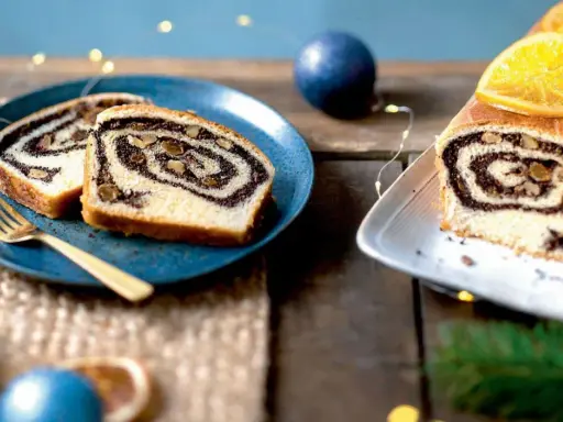 Tranches de roulé au pavot, garni de noix, sur une assiette bleue avec une fourchette dorée. Un autre roulé décoré d'orange est à droite.