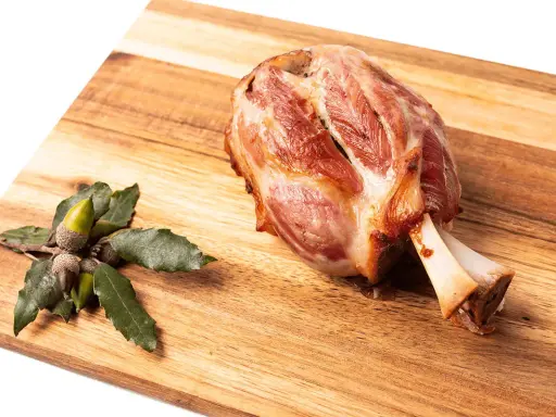 Codillo de cerdo asado, jugoso y dorado, reposa sobre una tabla de madera junto a unas bellotas.