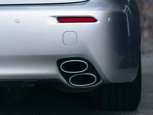 Doppio scarico cromato su un'auto sportiva argentata, parte del collettore di scarico. Sensori di parcheggio e fanale posteriore rosso visibili.