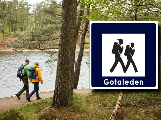 Två vandrare följer en stig vid en sjö, med en skylt för Gotaleden etapper.