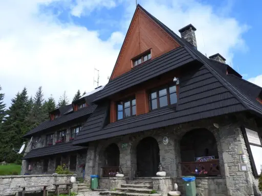 Kamienny schron turystyczny pod gorącymi szczytami, z drewnianą przybudówką i czarnym dachem, otoczony lasem świerkowym.