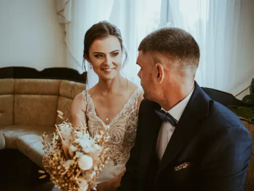Uśmiechnięta panna młoda w sukni z koronkami i pan młody w granatowym garniturze. W tle kanapa i zasłony. Krótka ceremonia, bo ile trwa ślub cywilny?