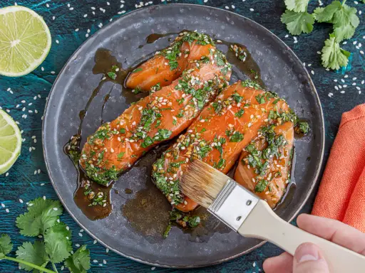 Lachsfilets werden mit einer asiatischen Marinade aus Sojasauce, Ingwer, Knoblauch, Kräutern und Sesam bestrichen. Dazu Limettenspalten und Koriander.