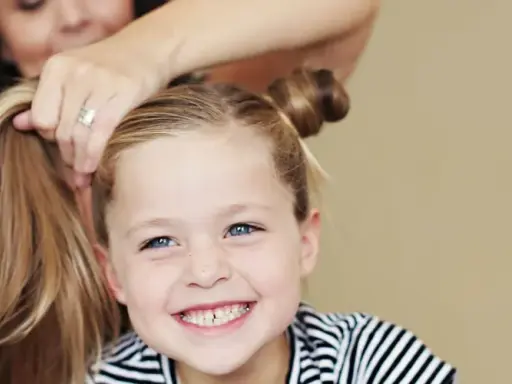 Ein Mädchen mit blauen Augen und Sommersprossen lächelt, während ihre Mutter ihr die Haare zu niedlichen Dutts frisiert. Tolle Ideen für kinder frisuren!