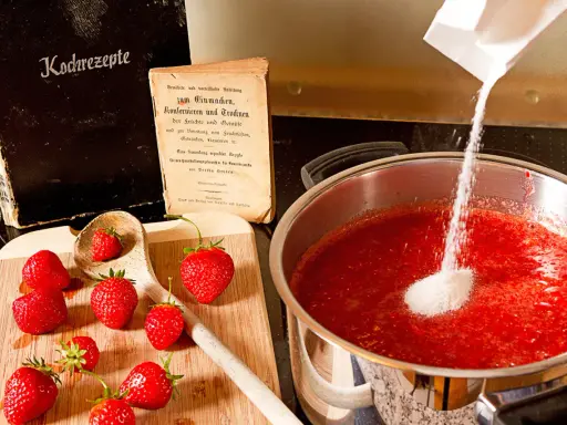 Zucker wird zu Erdbeeren in einem Topf gegeben, um zu erfahren, wann wird Marmelade fest. Alte Kochbücher liegen daneben.