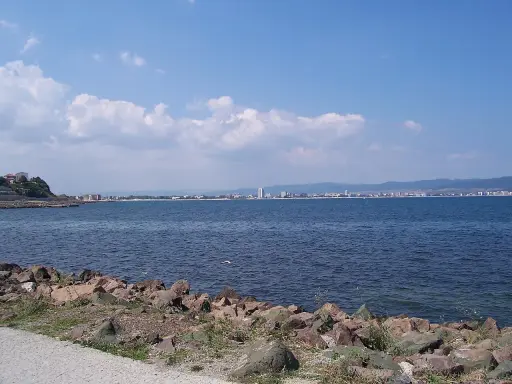 Słoneczny brzeg jak pić. Widok na morze i miasto w oddali, z kamienistym wybrzeżem na pierwszym planie.
